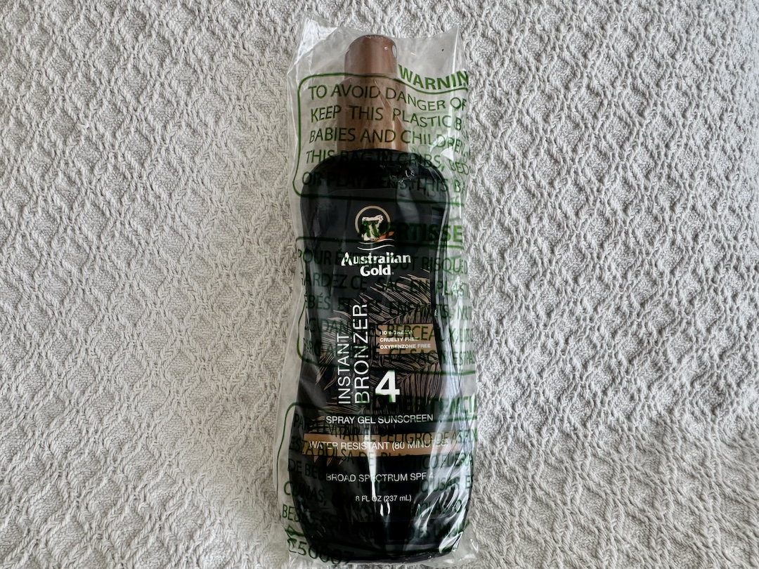 Image of a bottle of Australian Gold Spray Gel Sunscreen with Instant Bronzer SPF 4. The bottle is dark brown with a tropical-themed design, featuring the Australian Gold logo and a koala mascot. The label highlights the product's dual function as both a sunscreen and an instant bronzer, providing a sun-kissed glow while offering low-level sun protection. The spray gel is water-resistant, with details about the SPF and bronzing effects clearly visible on the front. The bottle is shown standing upright against a plain background.