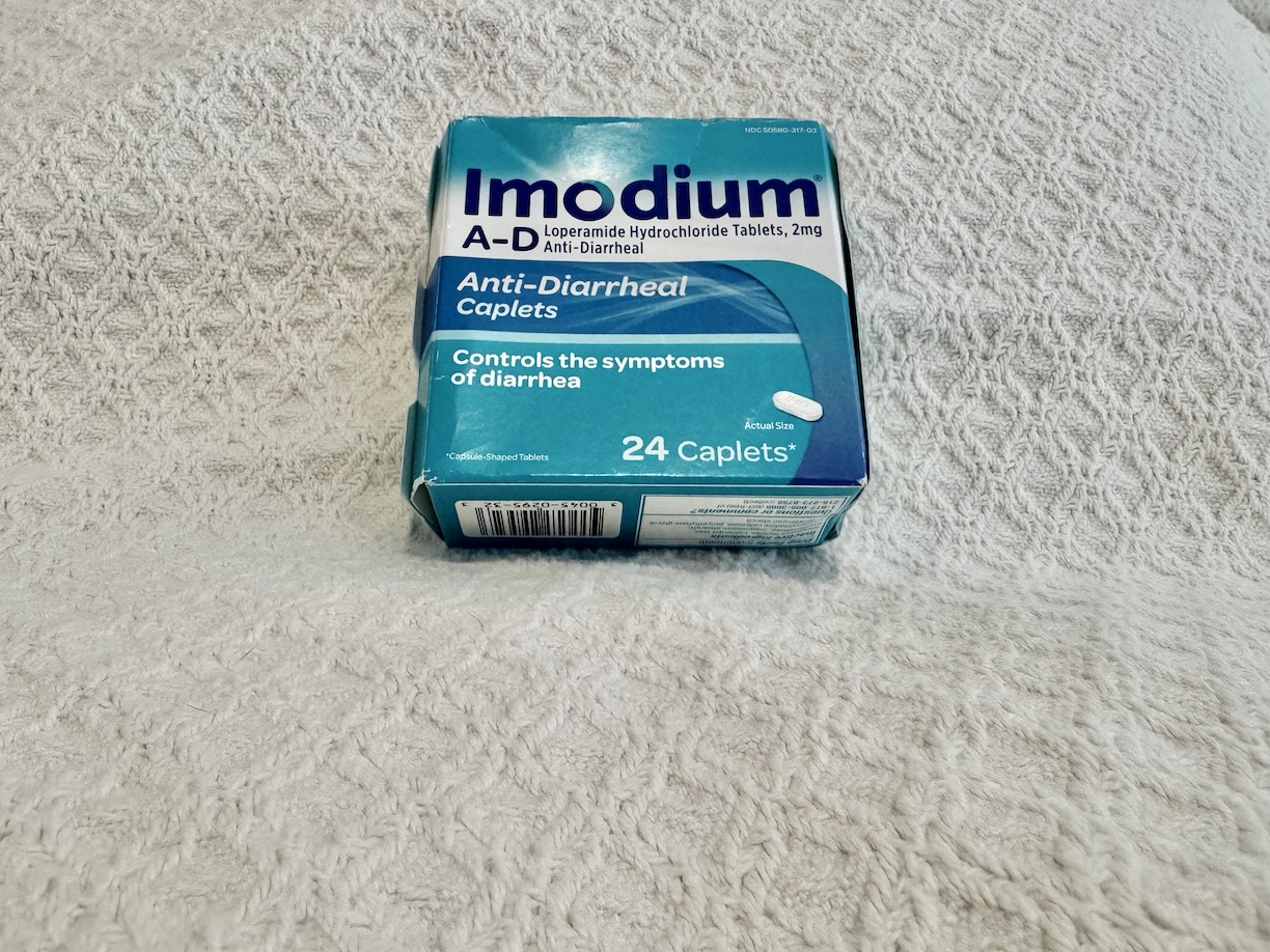 Close-up image of a box of Imodium A-D, an over-the-counter anti-diarrheal medication. The box is primarily blue with white text, featuring the brand name 'Imodium A-D' prominently at the top. The product description below highlights its use for controlling symptoms of diarrhea. The packaging also includes dosage information and key active ingredients. The box is positioned against a neutral background, making it easy to focus on the product details.