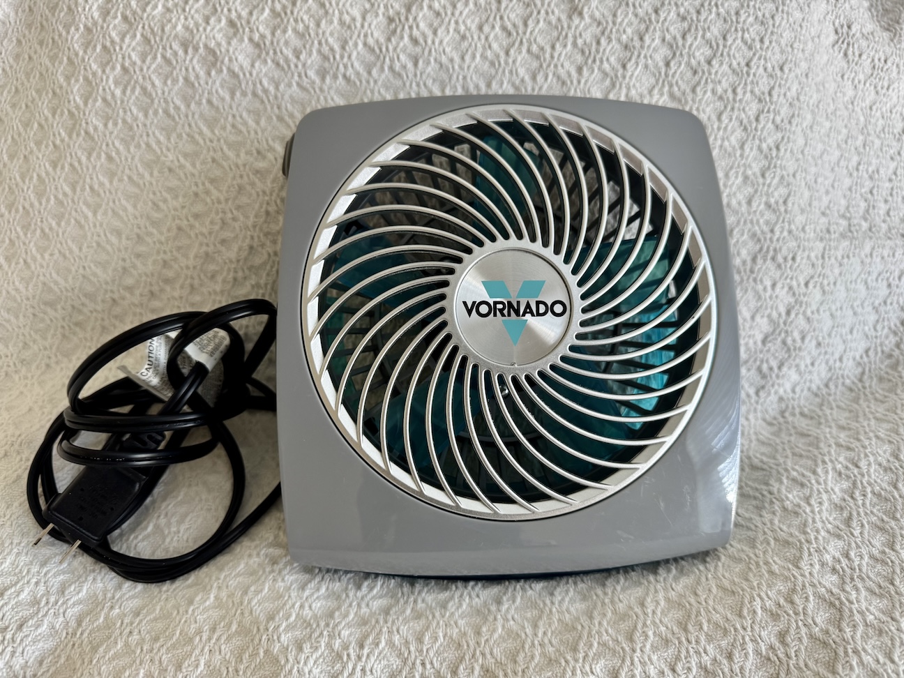 Image of a Vornado Fit Personal Fan, a compact and stylish desk fan designed for personal use. The fan has a sleek, modern design with a rounded base and a tilting head for adjustable airflow. It is available in a vibrant color, adding a pop of style to any workspace. The fan's front grille features concentric circles, and the control switch is easily accessible on the base. The fan is shown on a desk, highlighting its portability and suitability for cooling small personal spaces.